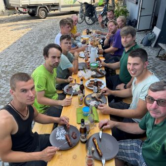 Das Team der "Grünen Oase" bei der Abschiedsfeier von Lukas, sie sitzen an einem Partytisch und es wird gegrilltes gegessen.