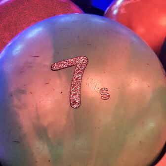 Bowlingkugel mit der Aufschrift „7s“ in Nahaufnahme, im Hintergrund verschwommen die Bowlingbahn im Neonlicht.