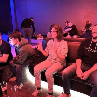 Vier Personen sitzen lachend auf einer Bowling-Couch, im Hintergrund spielen weitere Gruppen Bowling.